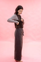 A woman modeling a dark brown vest over a gray long-sleeve shirt and dark brown wide-leg linen pants, standing against a solid pink studio background.