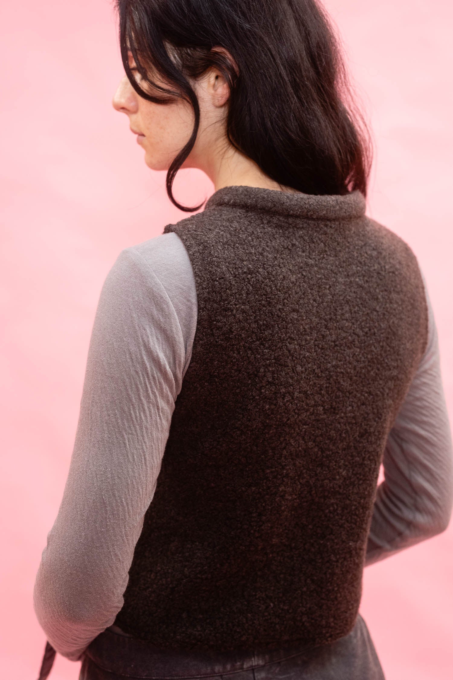 A woman shown from the back wearing a dark brown textured sleeveless sweater vest over a light gray long-sleeve top, set against a solid pink background.