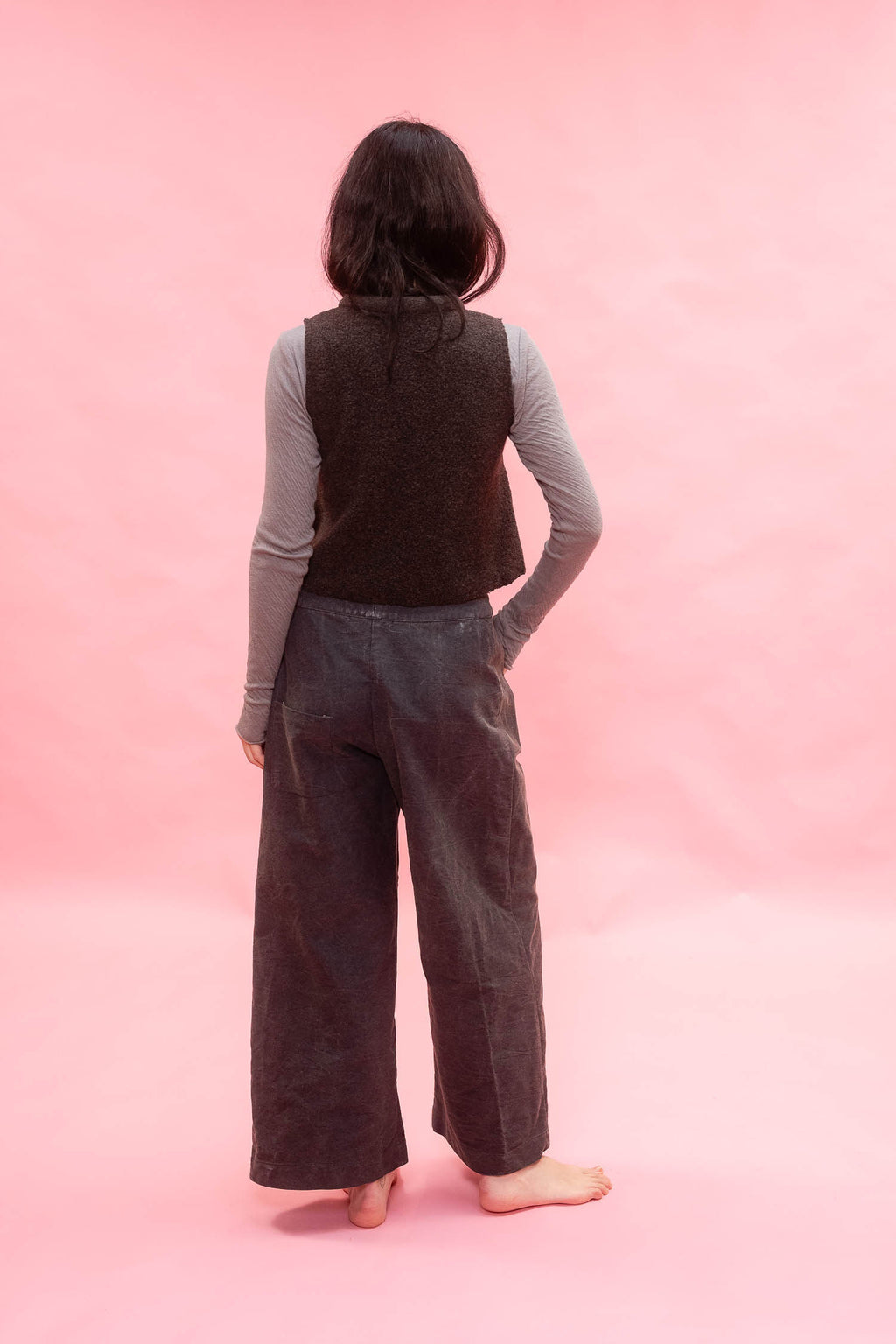 A person standing with their back to the camera, wearing a dark brown textured vest over a light gray long-sleeve shirt and dark wide-leg pants, set against a solid pink backdrop.