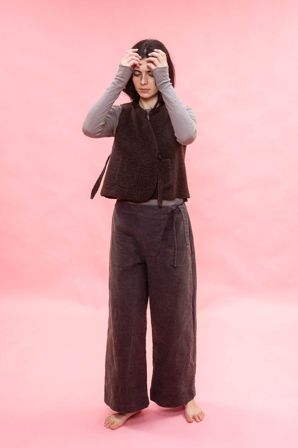 A person stands barefoot against a solid pink backdrop, wearing a dark brown sleeveless textured wool vest over a light gray long-sleeve top and dark brown wide-leg pants.