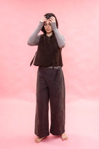 A person stands barefoot against a solid pink backdrop, wearing a dark brown sleeveless textured wool vest over a light gray long-sleeve top and dark brown wide-leg pants.