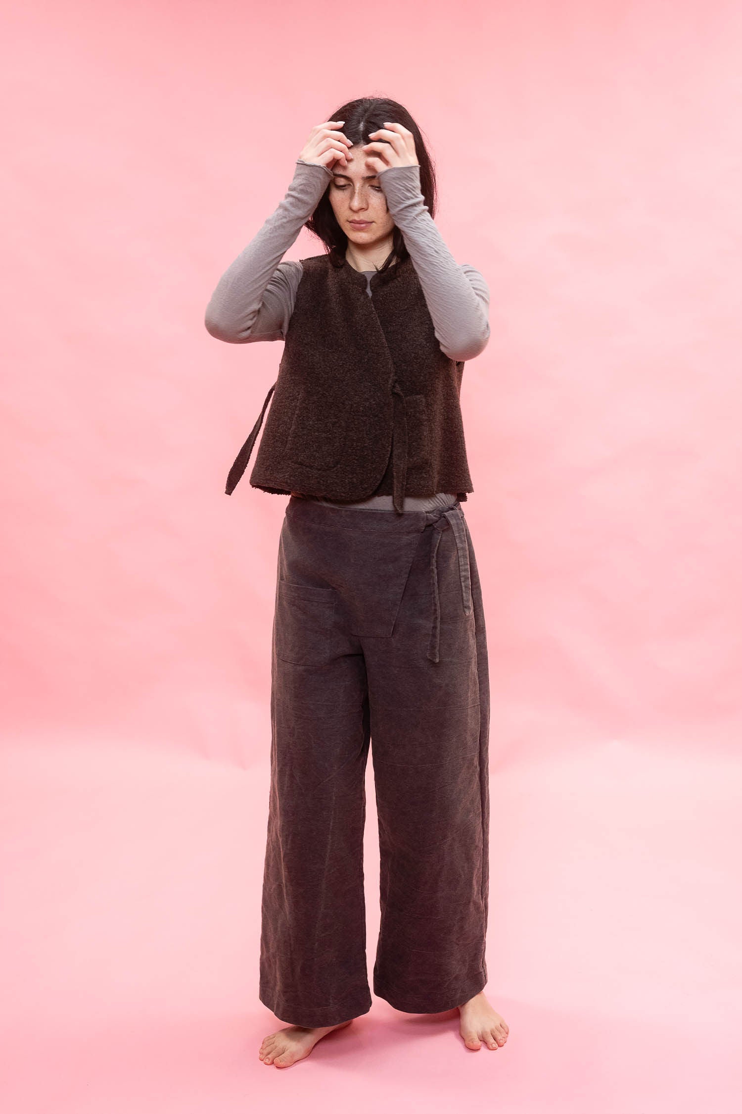 A person stands barefoot against a solid pink backdrop, wearing a dark brown sleeveless textured wool vest over a light gray long-sleeve top and dark brown wide-leg pants.