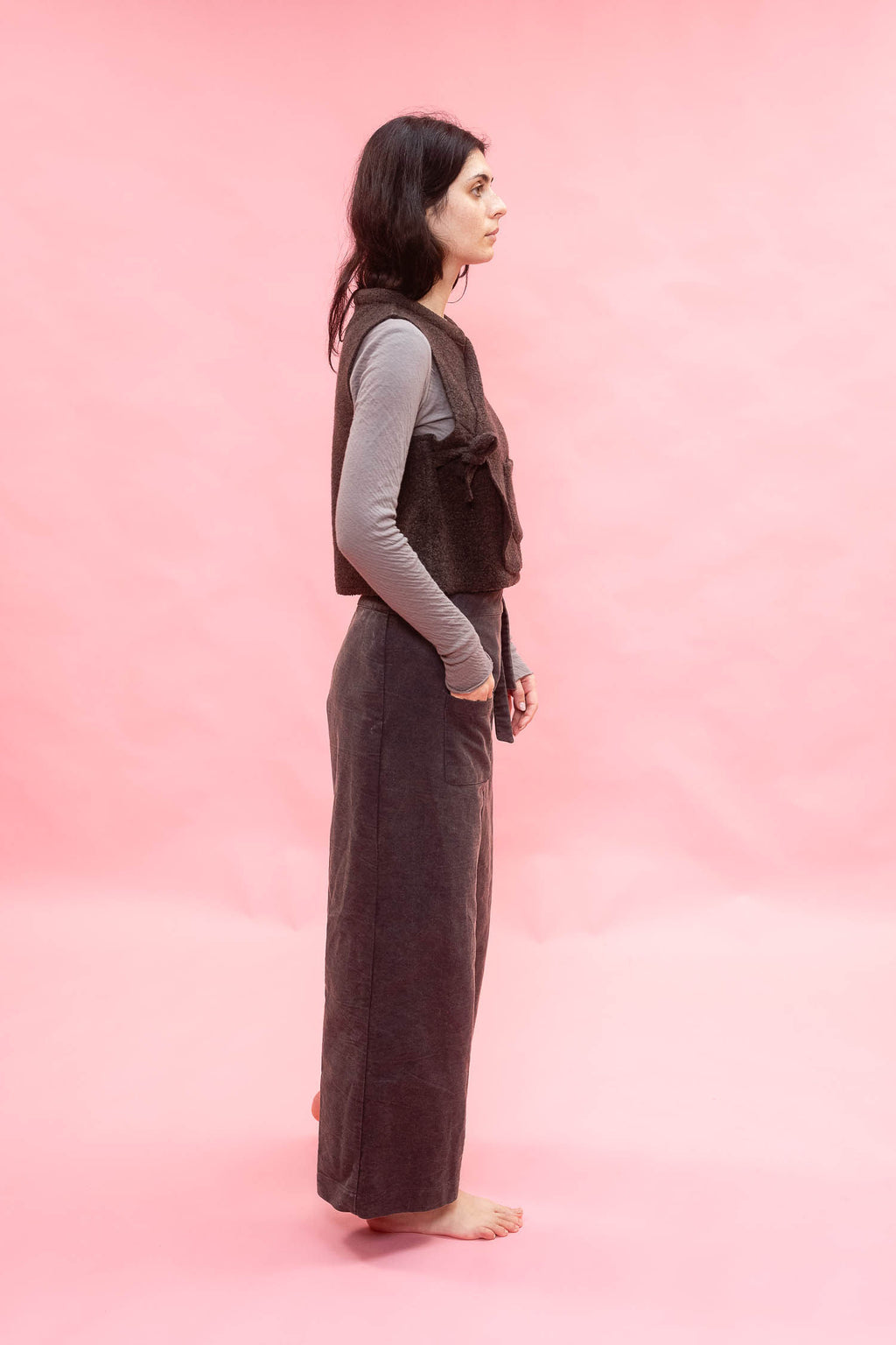 A woman stands in profile against a solid pink studio background, wearing a dark brown sleeveless vest over a light gray long-sleeve top and matching dark brown wide-leg pants.