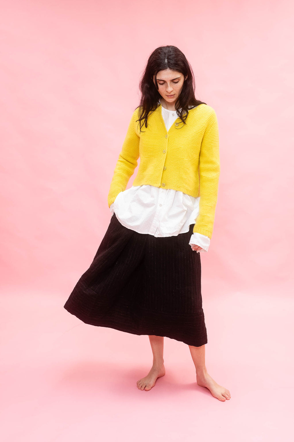 A woman stands barefoot against a solid pink studio background, wearing a bright yellow cardigan over a white top and a black wool pleated midi skirt.