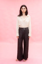 A woman stands barefoot against a solid pink studio background, wearing a cream long-sleeve blouse cotton Organdy with ruffled trim and dark brown wide-leg trousers.