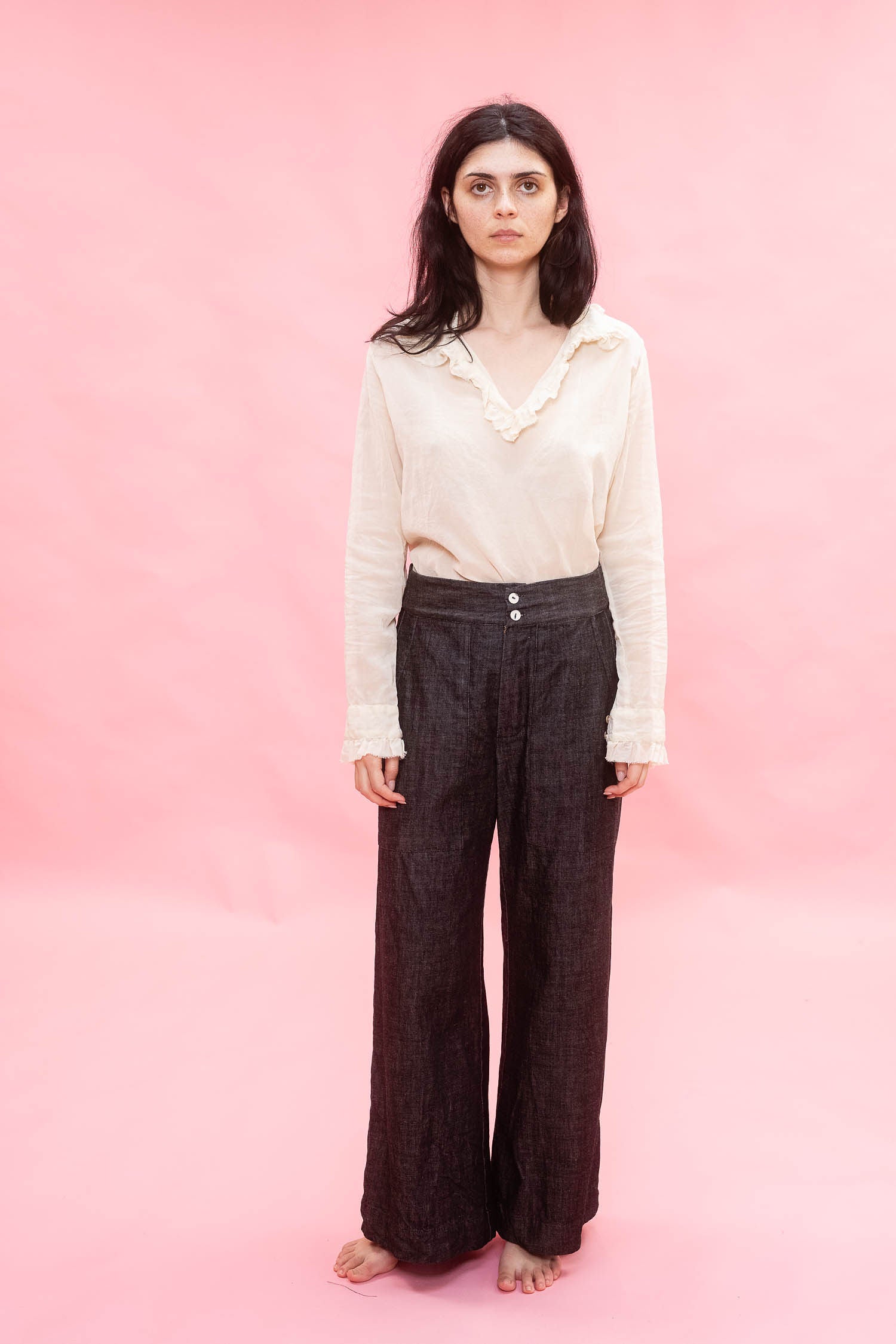 A woman stands barefoot against a solid pink studio background, wearing a cream long-sleeve blouse cotton Organdy with ruffled trim and dark brown wide-leg trousers.