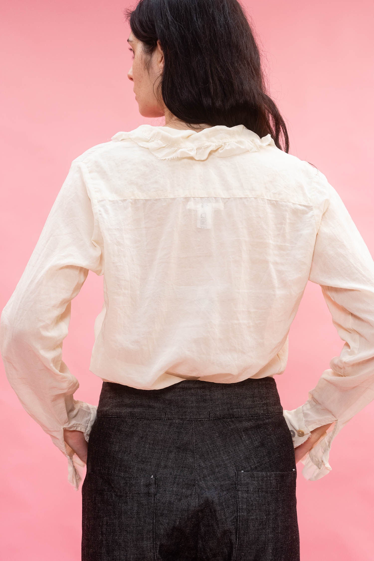 A woman shown from the back wearing a cream-colored, long-sleeved Japanese cotton organdy blouse with a ruffled collar and dark gray textured trousers, set against a solid pink background.
