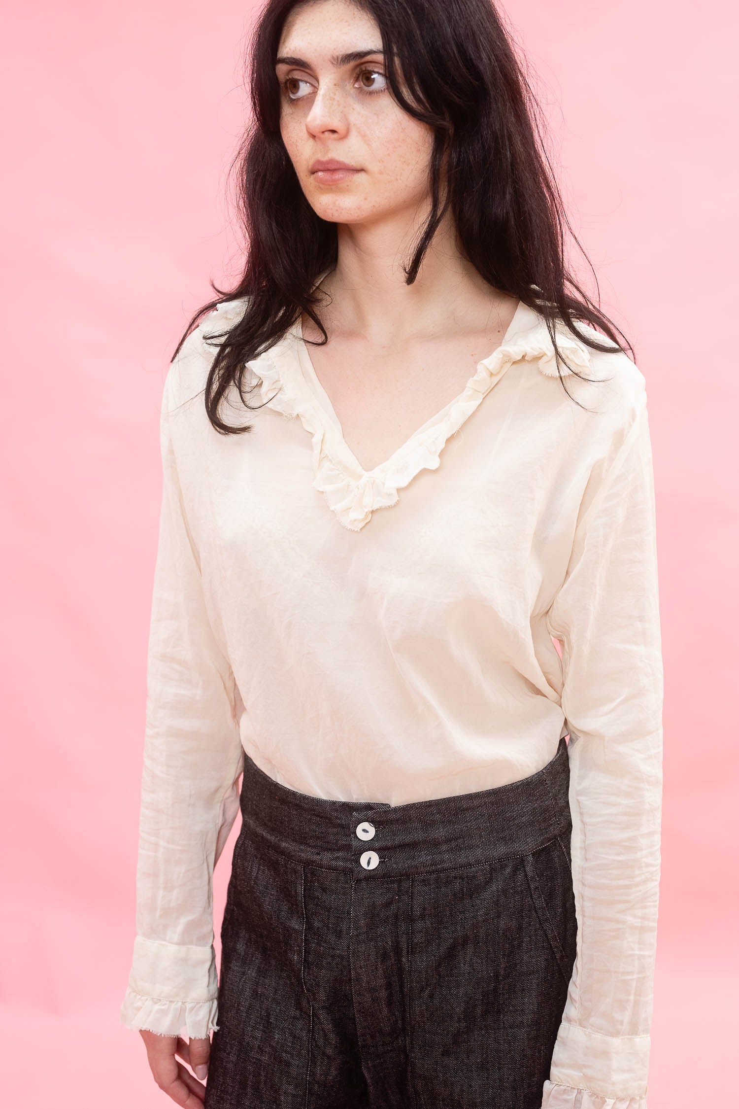 A woman wearing a cream-colored long-sleeve blouse with a ruffled v-neck collar and dark charcoal trousers, standing against a solid pink background.
