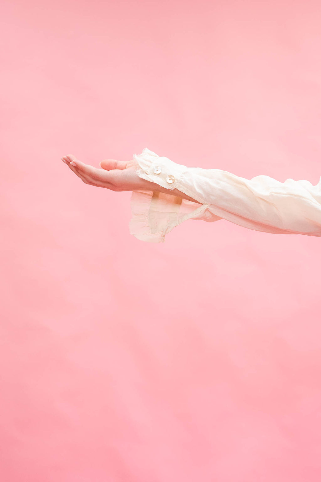 A person's outstretched hand and forearm wearing a sheer white long-sleeve blouse against a solid pink background.
