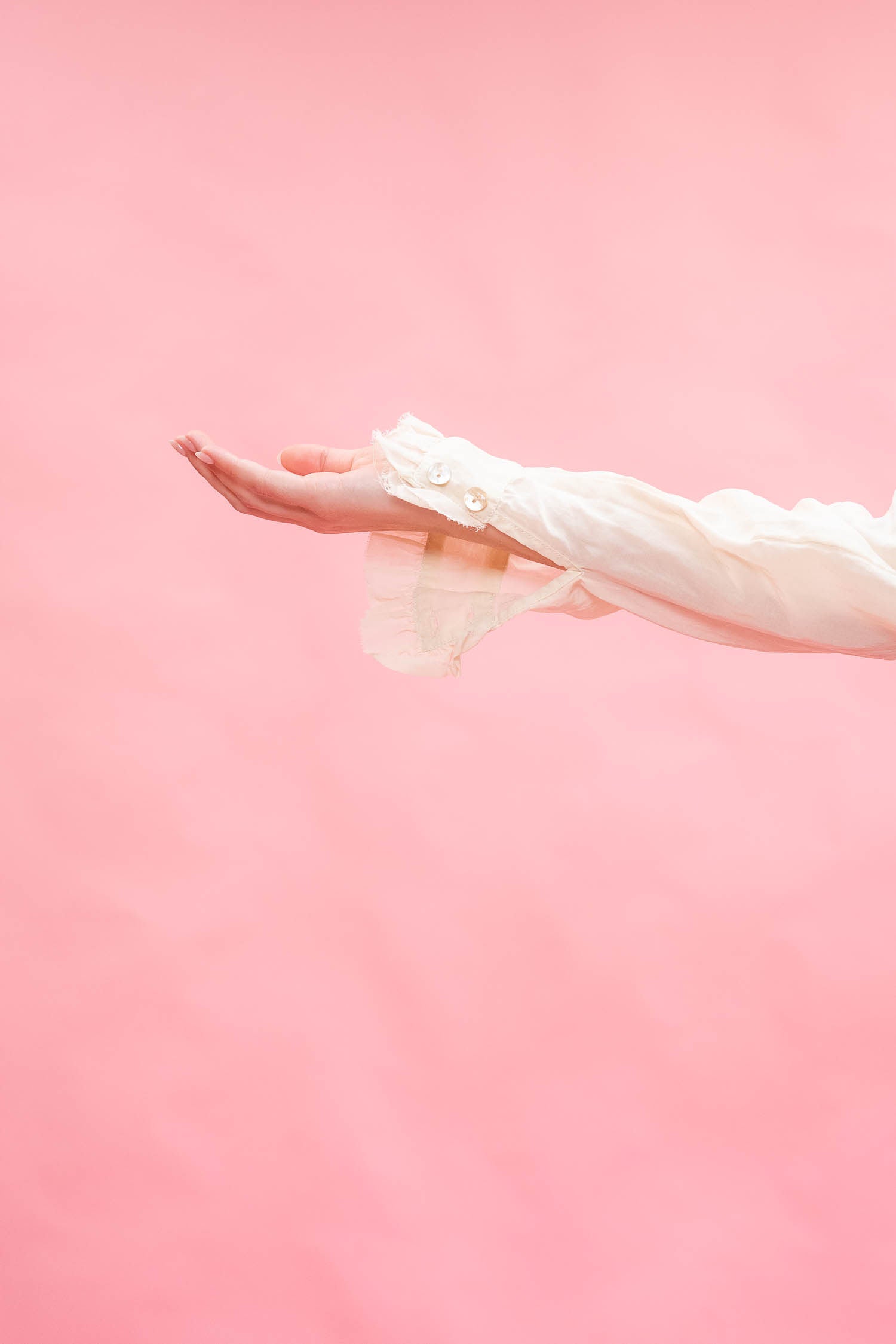 A person's outstretched hand and forearm wearing a sheer white long-sleeve blouse against a solid pink background.
