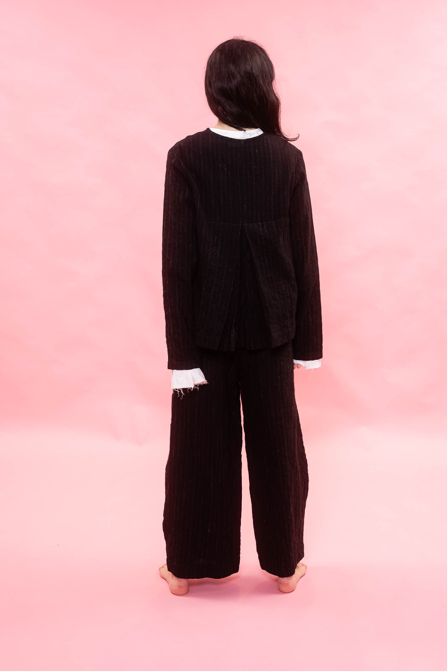 A model viewed from behind wearing a black pinstriped wool cropped jacket with a white collar and frayed cuffs blouse underneath, and matching wide-leg trousers, set against a solid pink background.