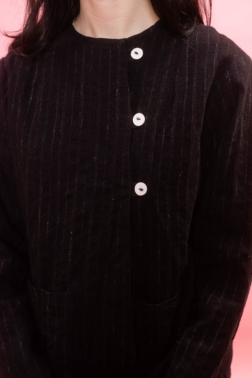A close-up of a person wearing a dark black wool pinstripe jacket with white buttons, set against a solid pink background.