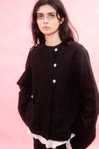 A woman wearing a black pinstripe wool button-front jacket over a white ruffled shirt, posed against a solid pink background.
