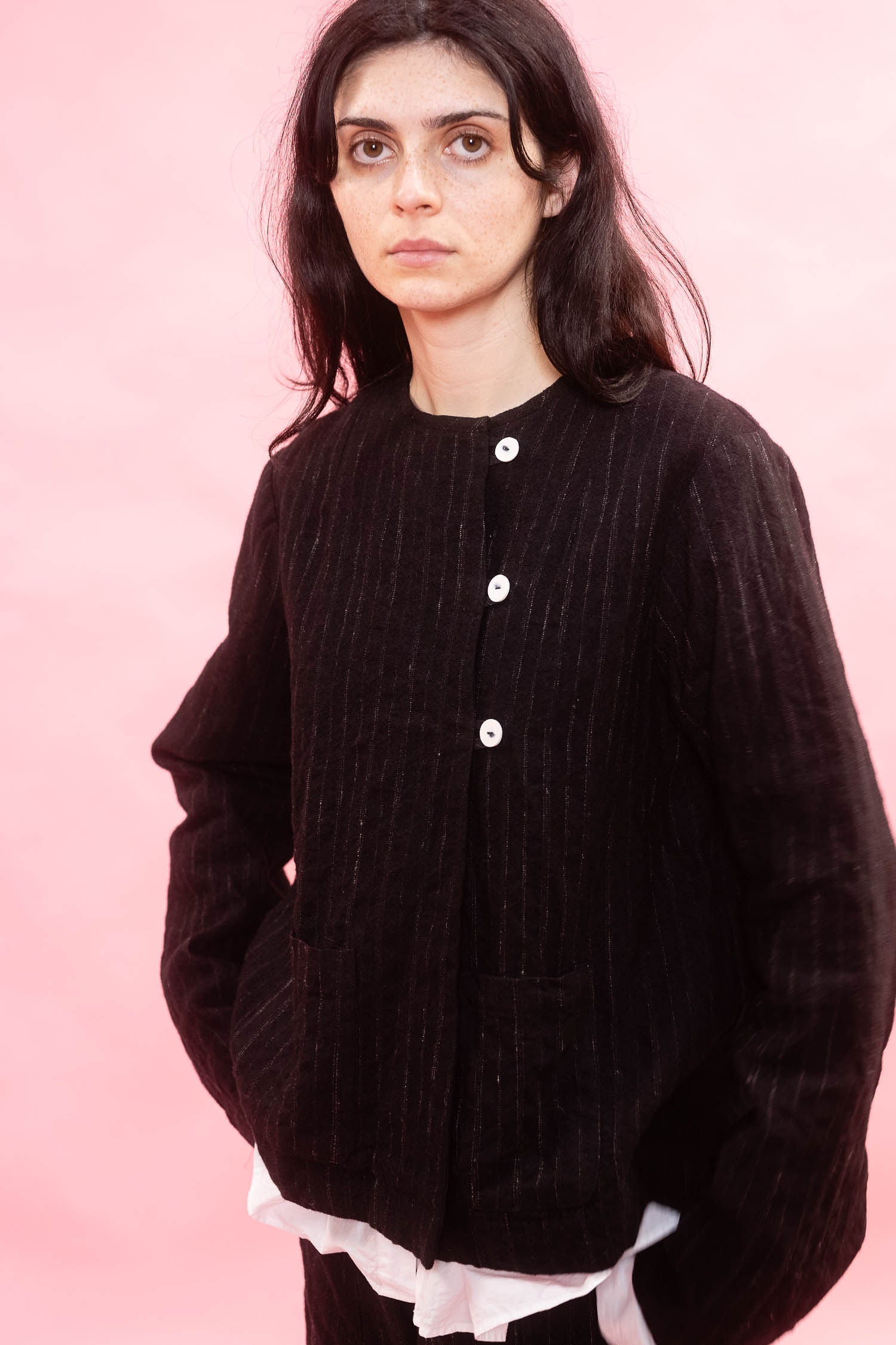 A woman wearing a black pinstripe wool button-front jacket over a white ruffled shirt, posed against a solid pink background.