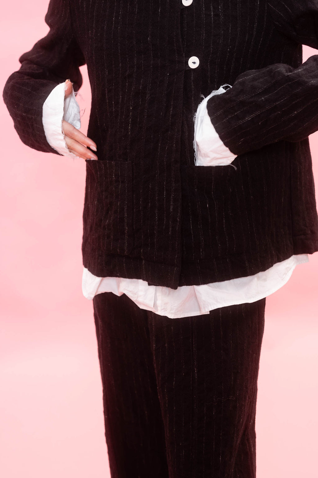A person wearing a dark black wool pinstripe jacket and matching pants, layered over a white ruffled shirt, against a solid pink background.