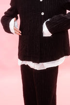A person wearing a dark black wool pinstripe jacket and matching pants, layered over a white ruffled shirt, against a solid pink background.