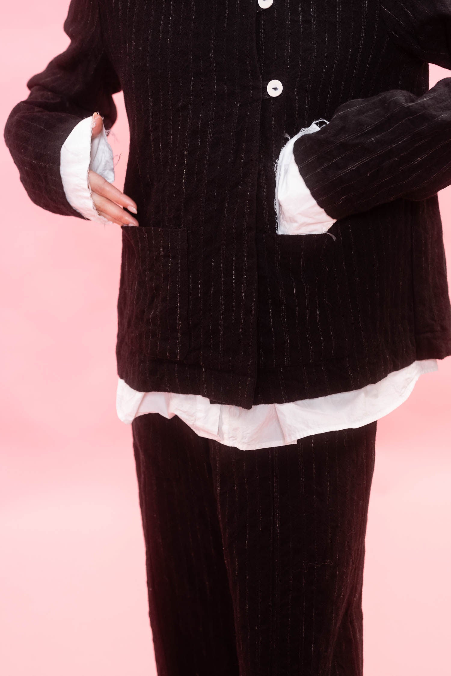 A person wearing a dark black wool pinstripe jacket and matching pants, layered over a white ruffled shirt, against a solid pink background.