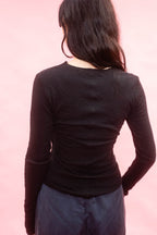 A person shown from the back wearing a fitted black long-sleeve tee, cut from Japanese cotton double jersey, and dark blue pants against a solid pink background.

