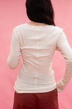 A person shown from the back wearing a fitted long-sleeve tee in a warm cream color, cut from Japanese cotton double jersey, and paired with dark reddish-brown pants, set against a solid pink studio background.
