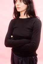 A woman with dark hair wearing a black long-sleeve crewneck tee, cut from Japanese cotton double jersey, standing with arms crossed against a solid pink background.
