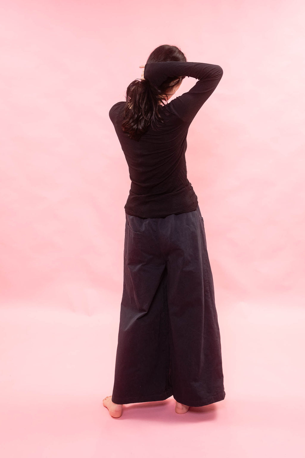 A woman viewed from behind wearing a black long-sleeve tee cut from Japanese cotton double jersey, and very wide-leg black pants, standing barefoot against a solid pink studio background.
