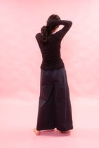A woman viewed from behind wearing a black long-sleeve tee cut from Japanese cotton double jersey, and very wide-leg black pants, standing barefoot against a solid pink studio background.
