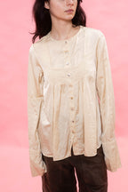 A model wearing a cream-colored, long-sleeved button-up blouse with a gathered pleated bodice, paired with dark brown pants, set against a solid pink background.