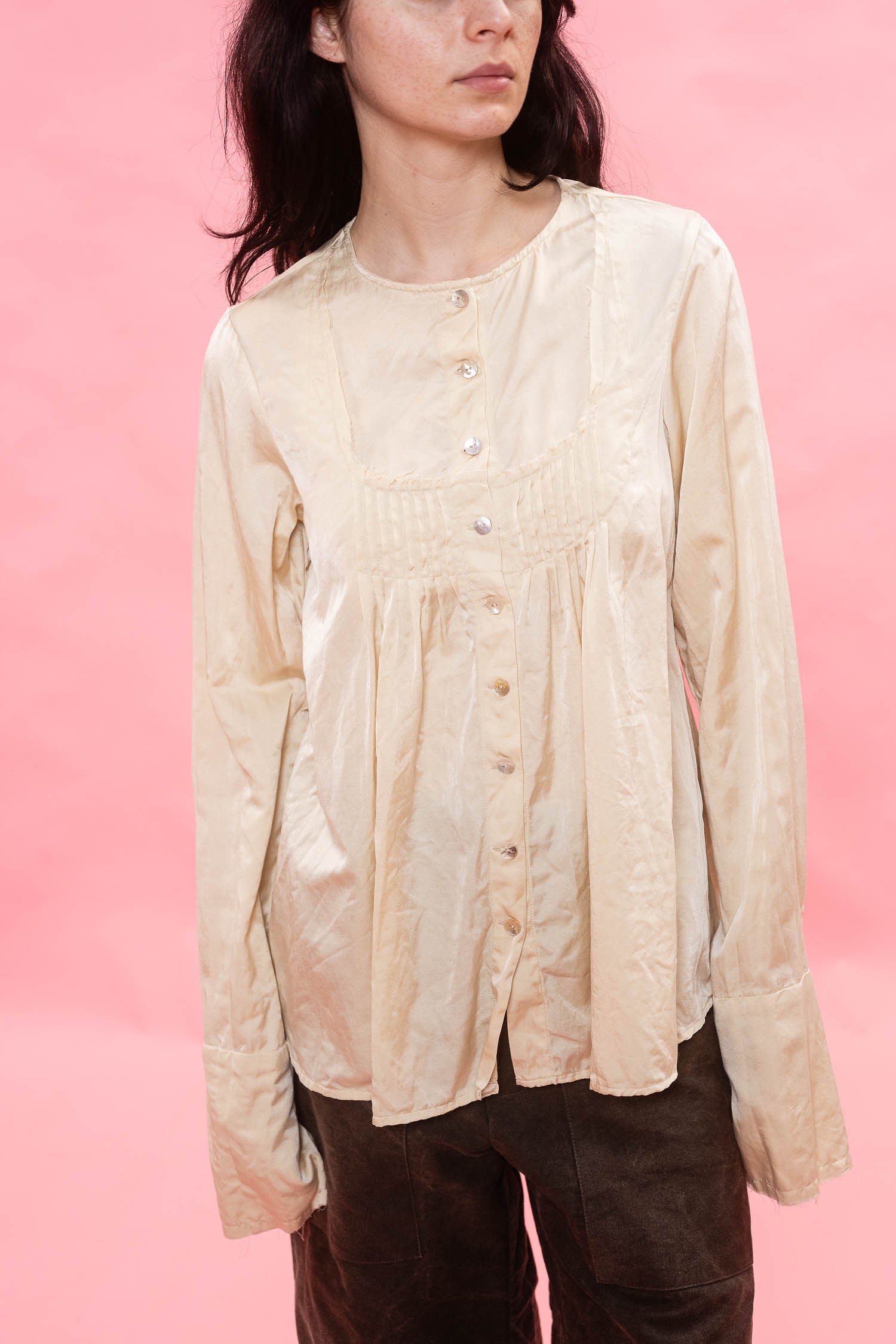 A model wearing a cream-colored, long-sleeved button-up blouse with a gathered pleated bodice, paired with dark brown pants, set against a solid pink background.
