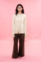 A woman stands against a solid pink studio background wearing a cream-colored, long-sleeved button-up cream pin tuck pleated silk cotton satin blouse and dark brown wide-leg pants.