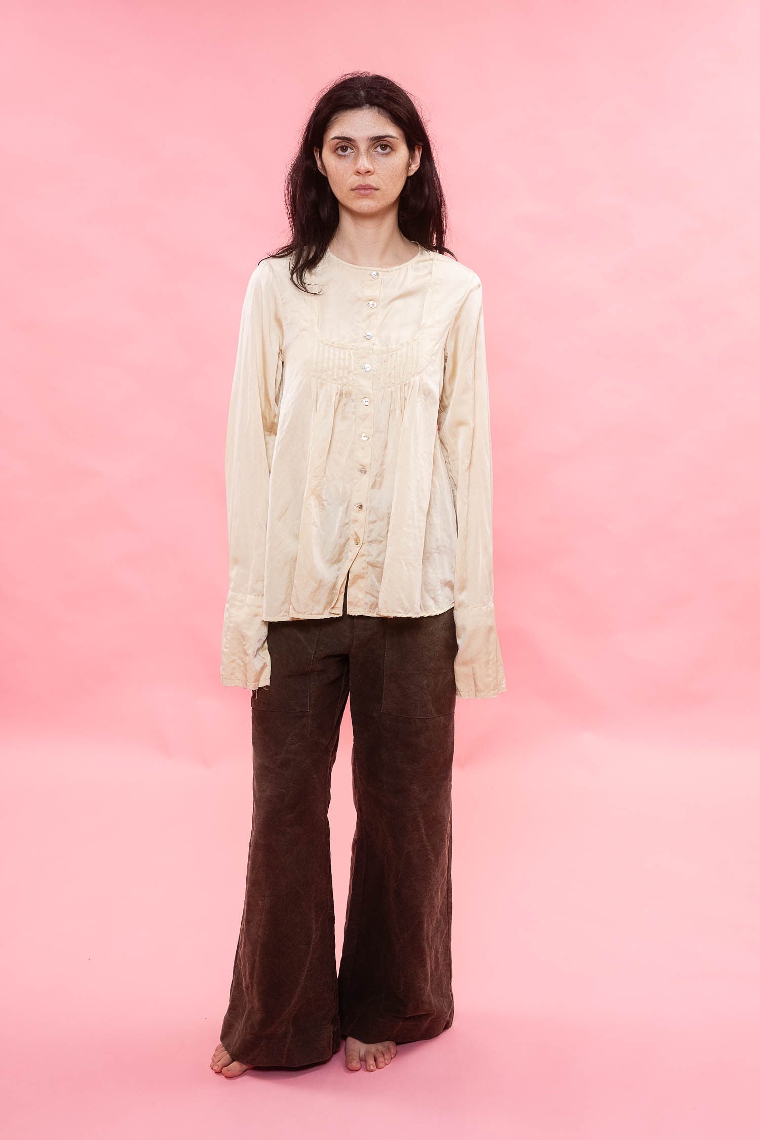 A woman stands against a solid pink studio background wearing a cream-colored, long-sleeved button-up cream pin tuck pleated silk cotton satin blouse and dark brown wide-leg pants.