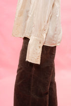 A close-up of a person wearing a cream-colored, satin long-sleeve blouse with raw, frayed edges and a dark brown  pants against a solid pink background.