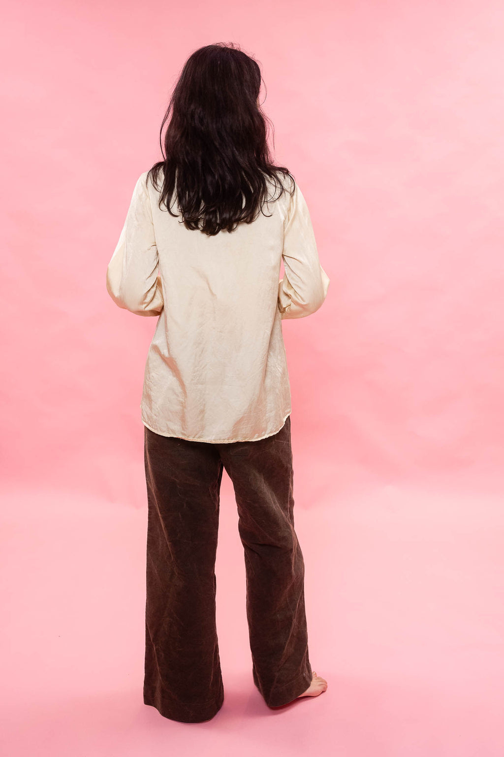 A person standing with their back to the camera, wearing a cream pin tuck pleated silk cotton satin and dark brown wide-leg pants, set against a solid pink background.