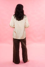 A person standing with their back to the camera, wearing a cream pin tuck pleated silk cotton satin and dark brown wide-leg pants, set against a solid pink background.