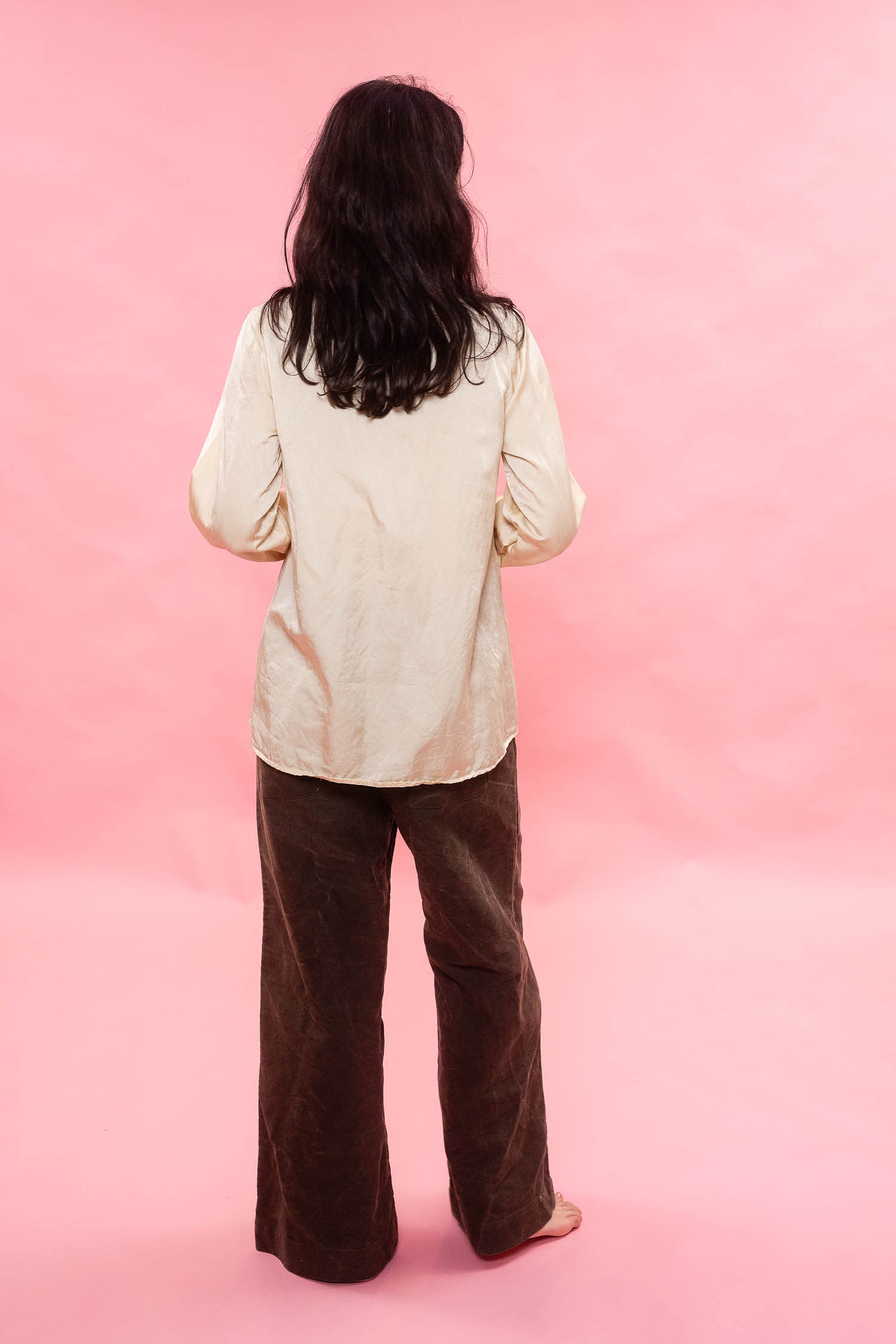 A person standing with their back to the camera, wearing a cream pin tuck pleated silk cotton satin and dark brown wide-leg pants, set against a solid pink background.