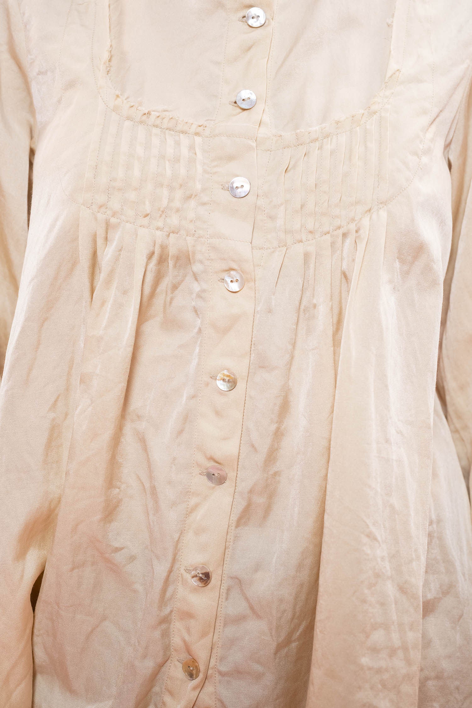 Close-up of a l cream-colored long-sleeve , satin blouse with a pleated bodice, decorative neckline, and a vertical button placket with white buttons.