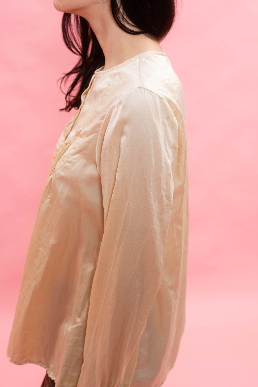 A woman wearing a loose, long-sleeve cream-colored blouse with a subtle sheen, shown in profile against a solid pink background.