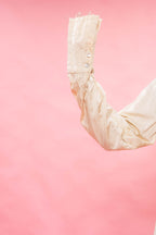 A close-up of a cream satin or silk long sleeve with decorative silver buttons on the cuff, set against a solid pink background.