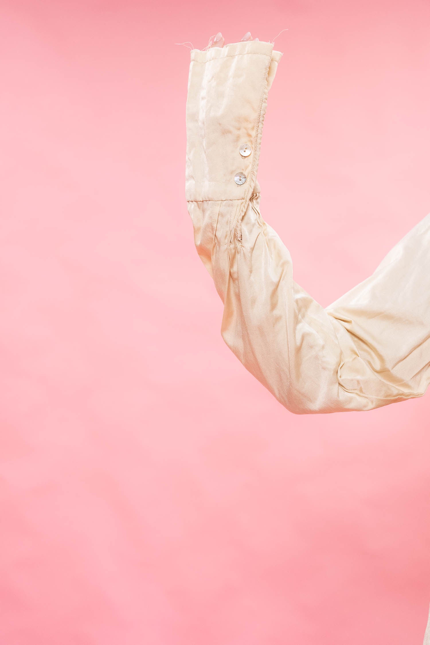 A close-up of a cream satin or silk long sleeve with decorative silver buttons on the cuff, set against a solid pink background.