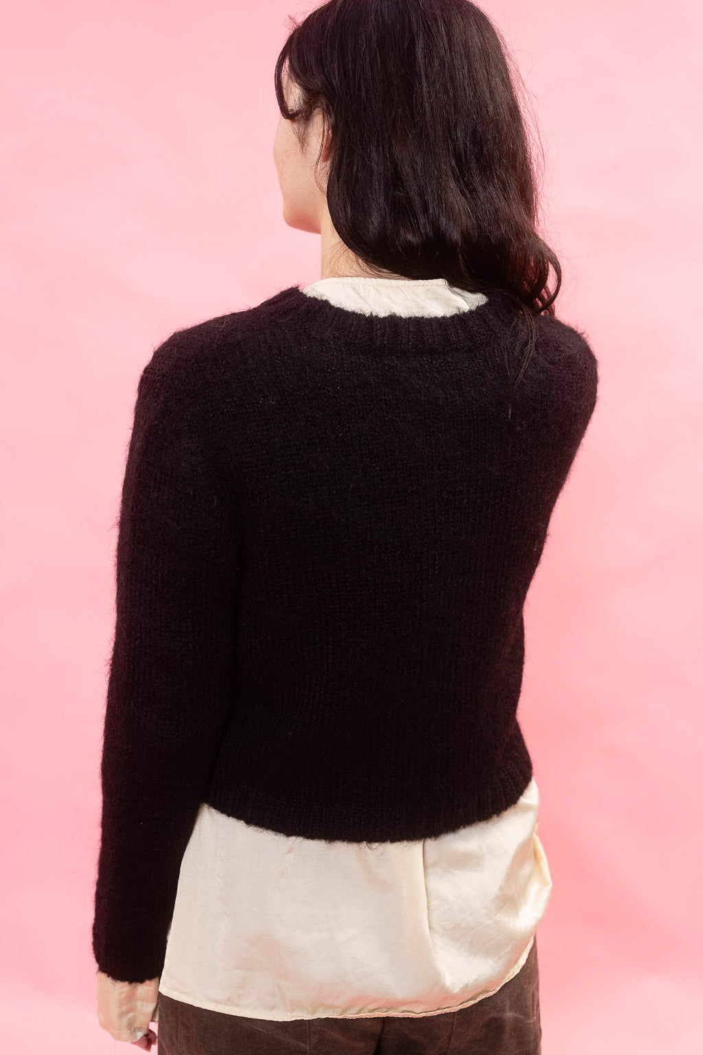 A woman shown from the back wearing a black fluffy knit alpaca cardigan over a cream shirt, set against a solid pink background.
