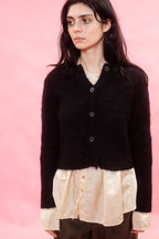 A woman wearing a black fluffy alpaca knit cardigan over a cream shirt and brown pants, standing against a solid pink background.
