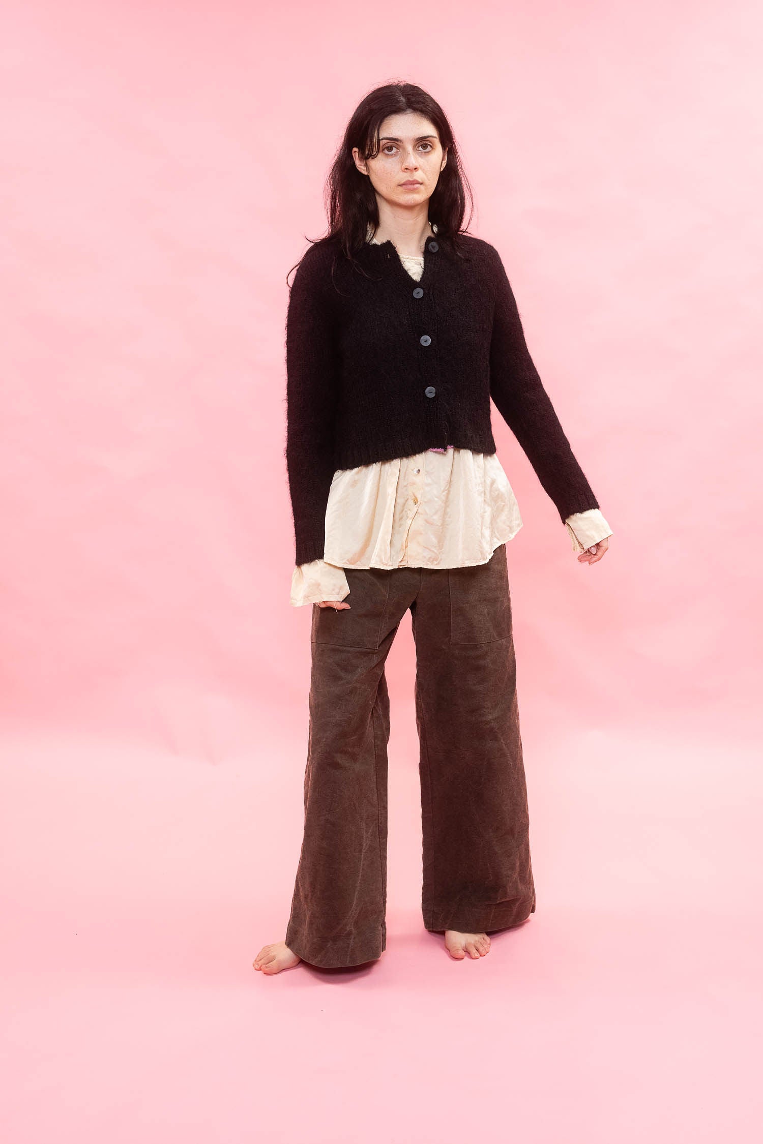 A woman stands barefoot against a solid pink studio backdrop, wearing a black cropped button-front alpacacardigan over a cream ruffled blouse and dark brown wide-leg pants.

