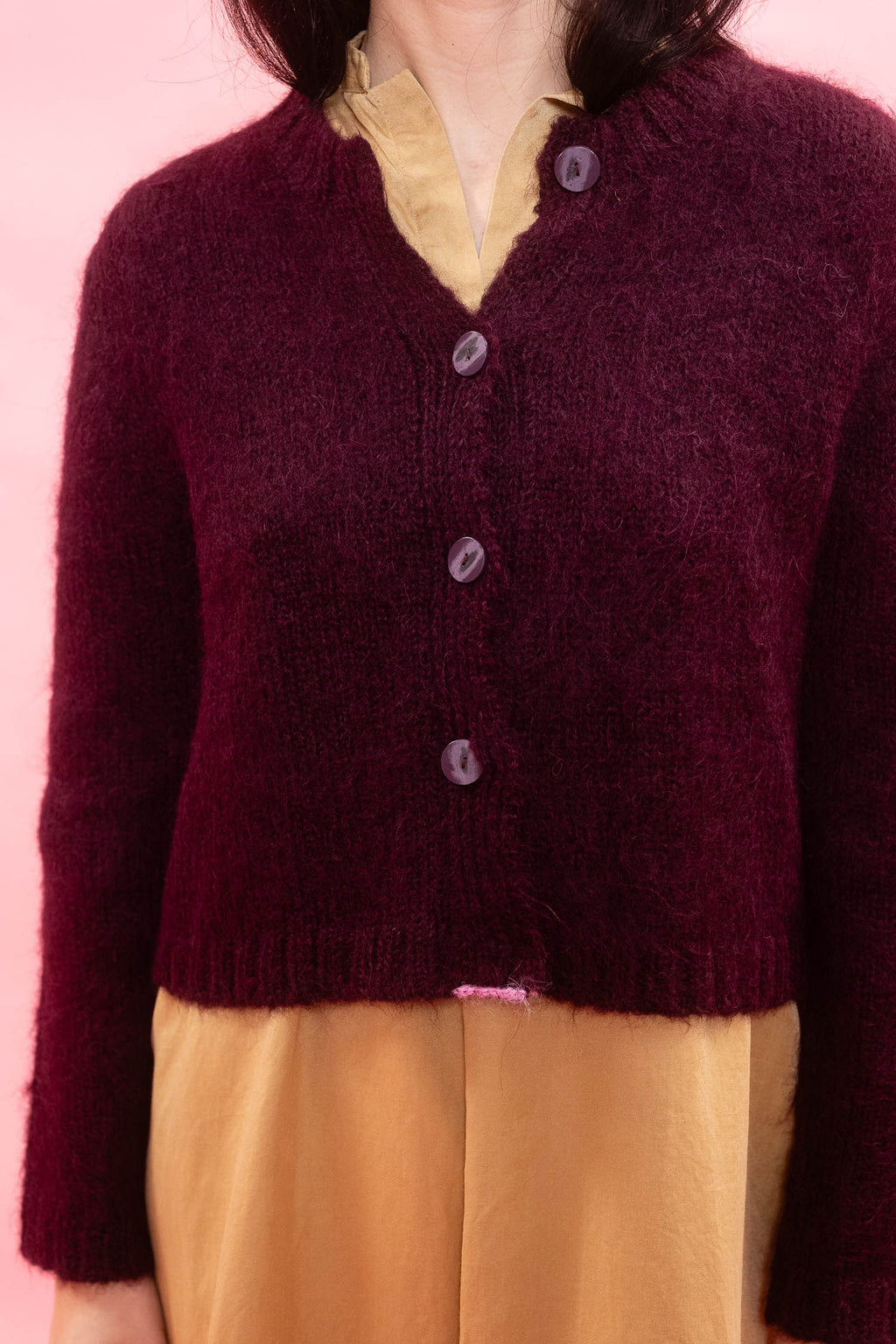 A close-up of a person wearing a fluffy knit alpaca cardigan in deep plum over a mustard yellow dress, set against a solid pink background.
