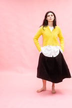 Woman wearing a yellow cashmere hand knit cardigan, white blouse, and black wool linen skirt on a pink background