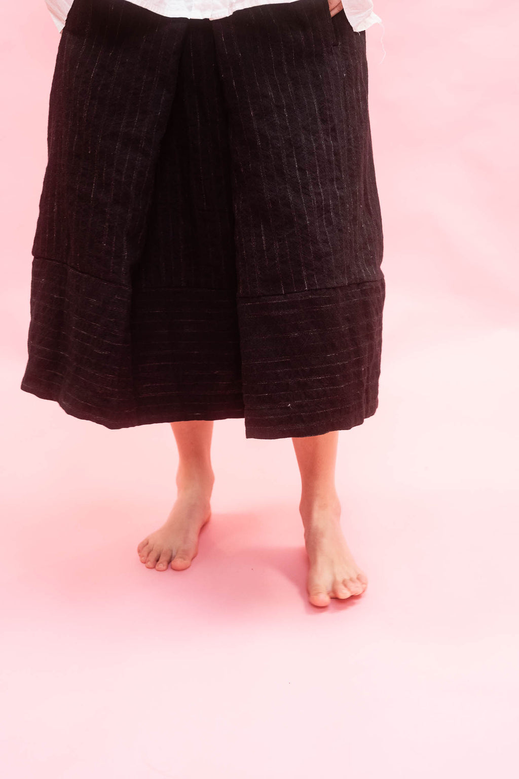 A person wearing a black textured midi skirt, shown from the waist down against a solid pink background.