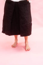 A person wearing a black textured midi skirt, shown from the waist down against a solid pink background.