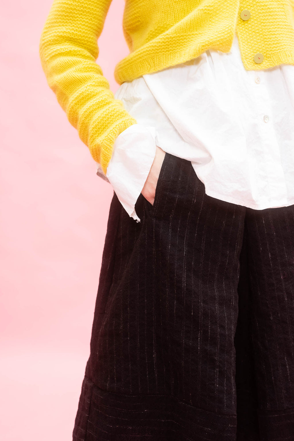 A close-up of a person wearing a bright yellow knit cardigan over a white button-up shirt and black pinstriped wool skirt, set against a solid pink background.