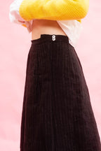 A close-up of a person wearing a black wool pinstriped skirt with a high waist and wood button detail, styled with a white shirt and yellow sweater against a solid pink background.