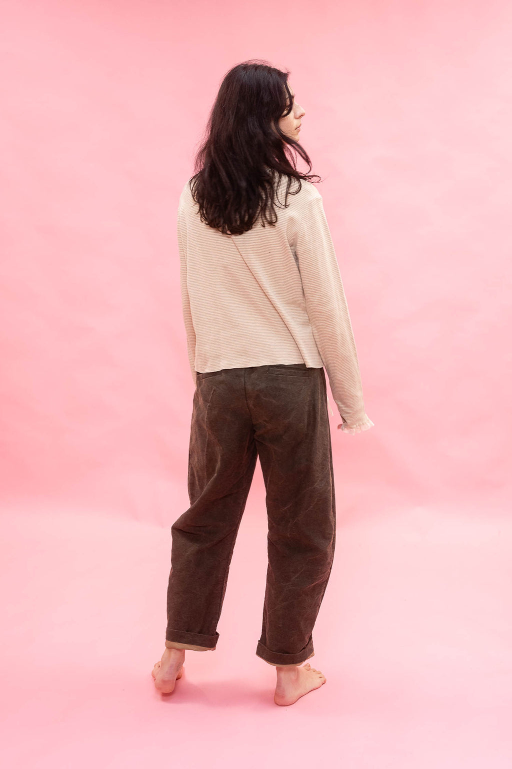 A woman is shown from behind wearing a light beige long-sleeve shirt and dirt green cotton patina pants, standing barefoot against a solid pink studio backdrop.