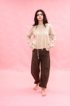 A woman stands barefoot against a solid pink studio backdrop, wearing a light beige long-sleeve knit top with white silk ribbon detail and dirt green cotton patina pants.