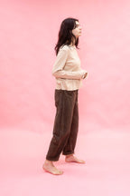 A woman stands in profile against a solid pink studio backdrop, wearing a light beige long-sleeve top and dirt green cotton patina pants.